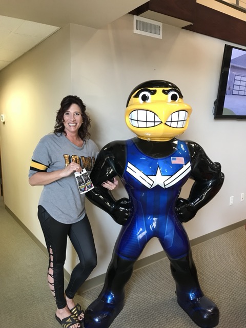 Brown haired woman dressed in gray tshirt, blue jeans, black sandals standing next to Iowa Herky wearing blue singlet wrestling statue